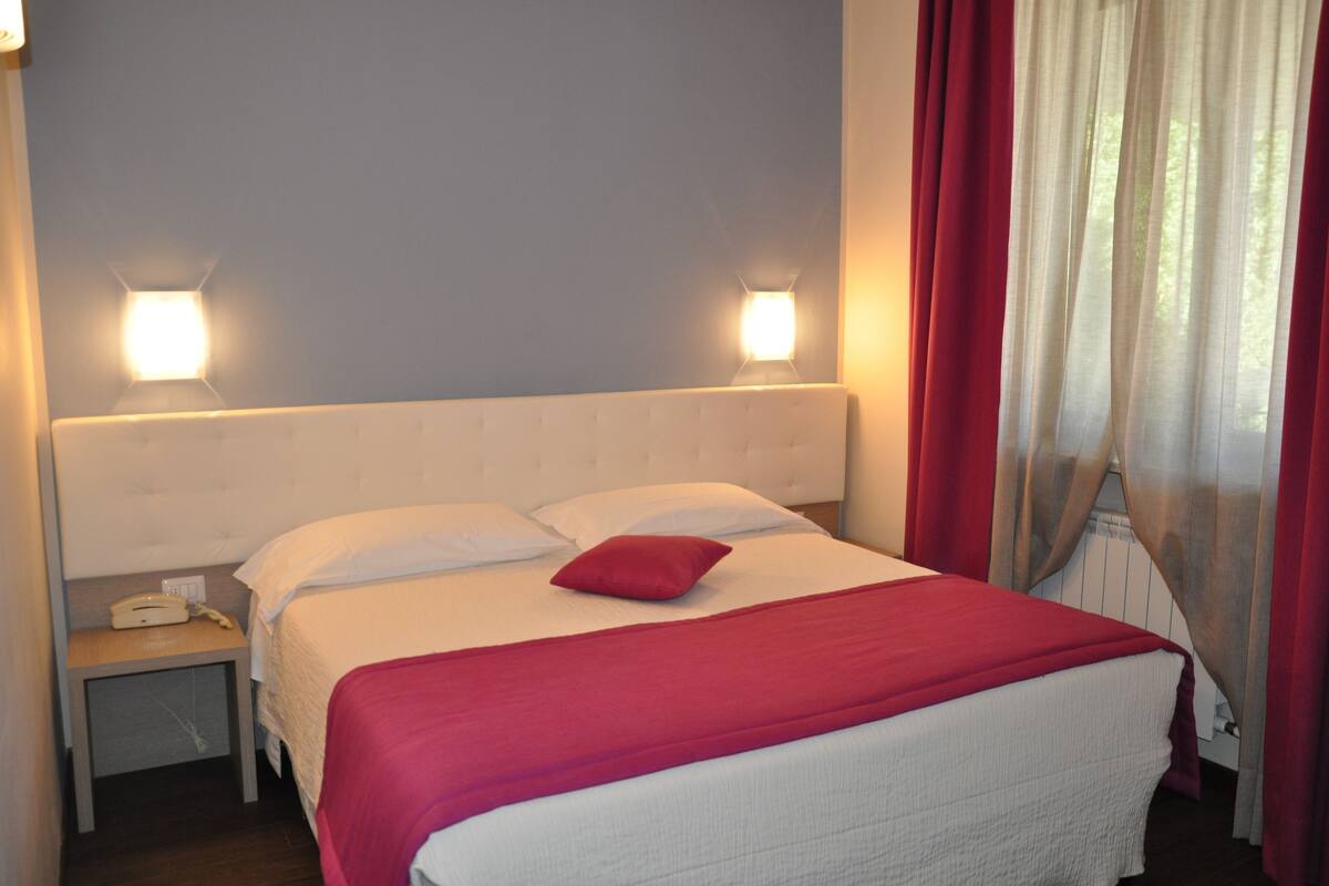 Ein ordentlich bezogenes Bett mit roter Decke und Kissen, ein Nachttisch mit Telefon sowie zwei an der Wand befestigte Lampen.