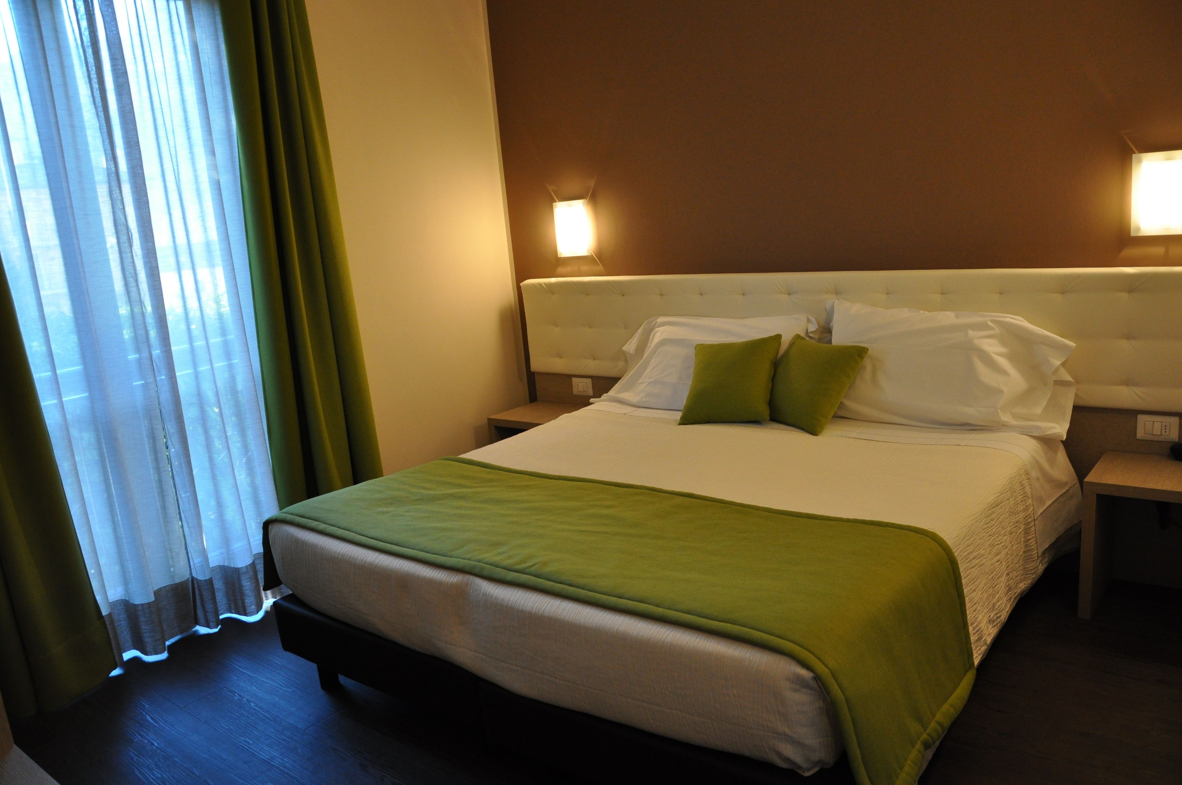 A neatly made bed with a green bedspread and pillows, a bedside table with a lamp, and a window with sheer curtains.