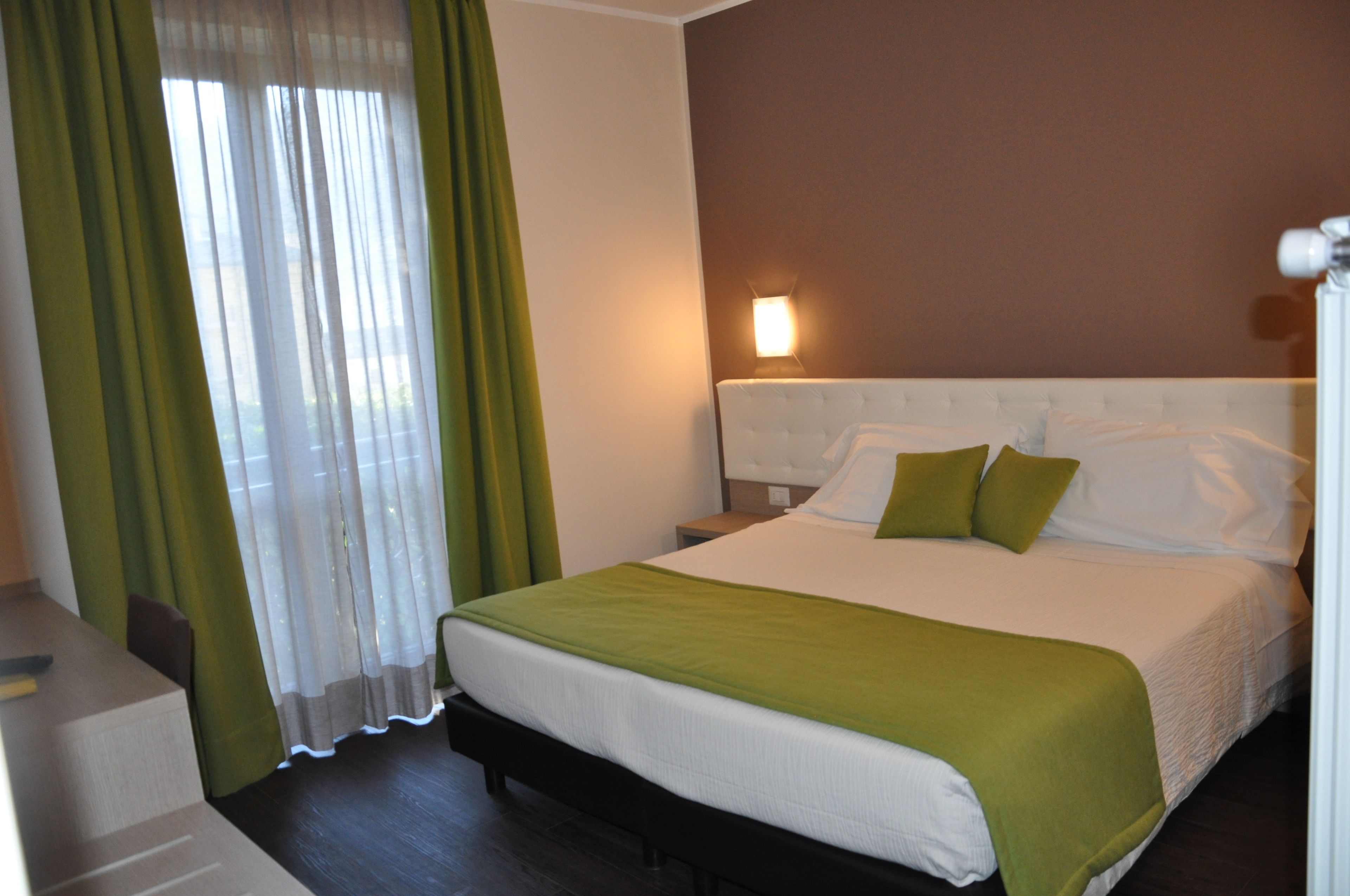 A hotel room with a bed, green curtains, a nightstand, and a wall-mounted lamp.