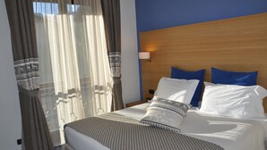 A hotel room with a bed, curtains, a nightstand, and a wall-mounted lamp. - Saint Pierre (Saint-Pierre)
