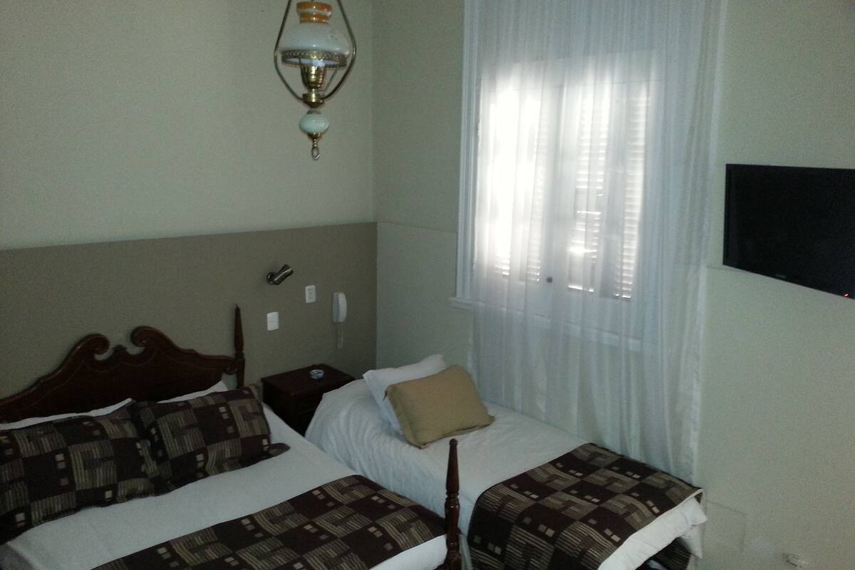Una cama doble con ropa de cama estampada, una mesita de noche con lámpara y una ventana con cortinas traslúcidas.