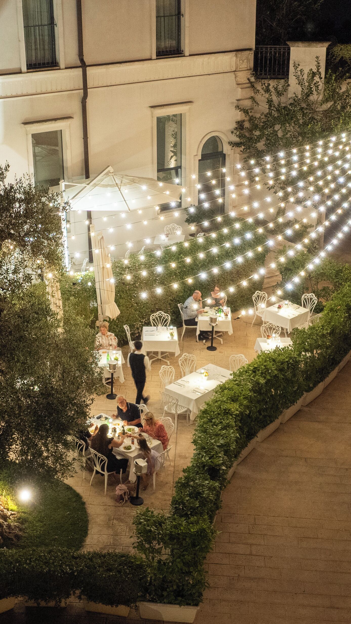An outdoor dining area with tables and chairs, surrounded by greenery and string lights.