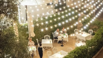 An outdoor dining area with tables and chairs, surrounded by greenery and string lights.