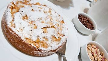 Un pastel redondo cubierto de azúcar glas sobre un plato blanco, acompañado de tazones de cereal y tazas de café.