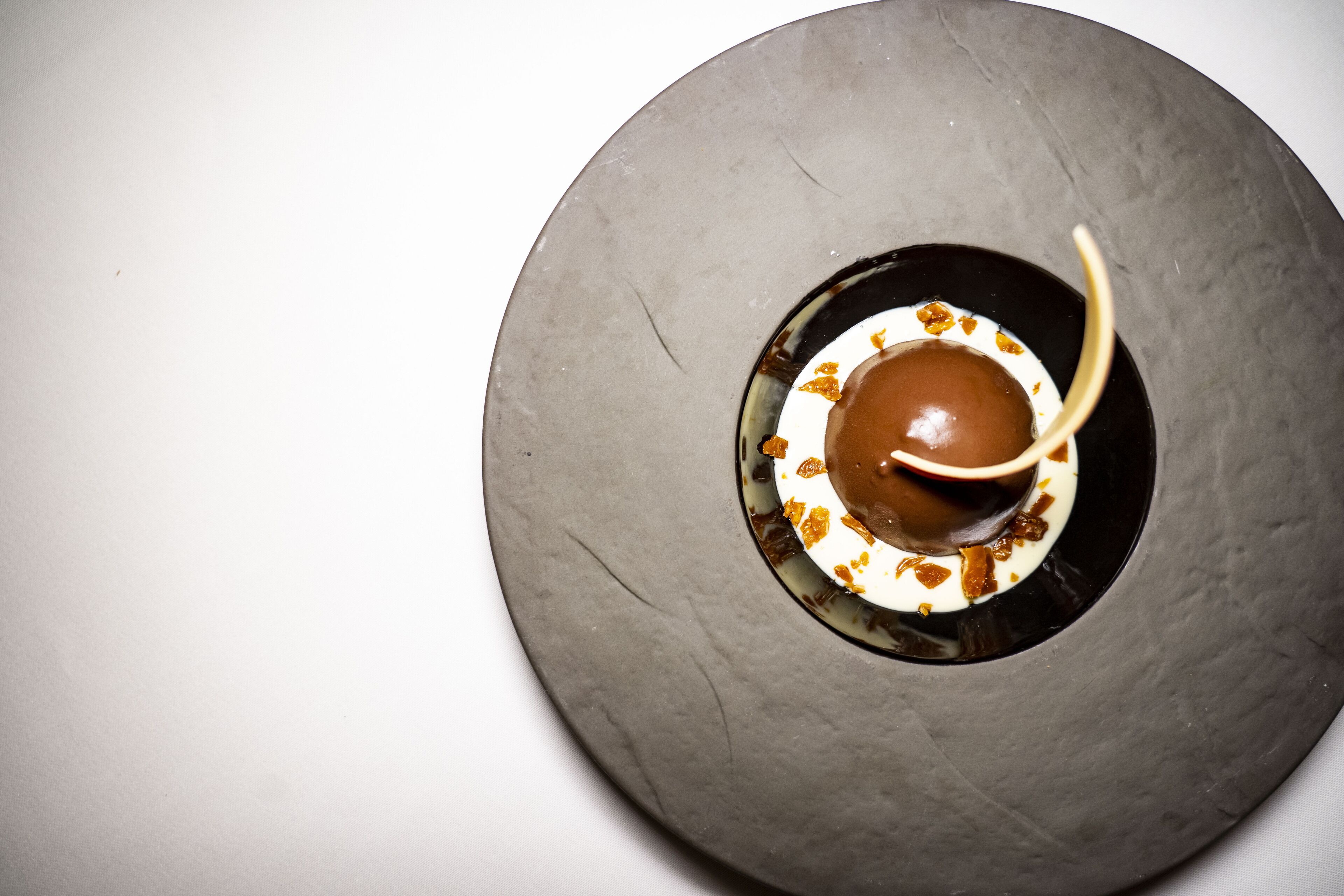 Ein runder, strukturierter Tisch mit einer Mittelloch, ein Schokoladendessert mit Karamellsauce und ein weißer Teller mit goldenen Flocken.