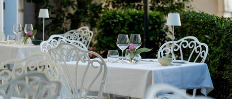 Un área de comedor al aire libre con sillas y mesas de metal blanco, cada conjunto con copas de vino y un pequeño jarrón con flores.