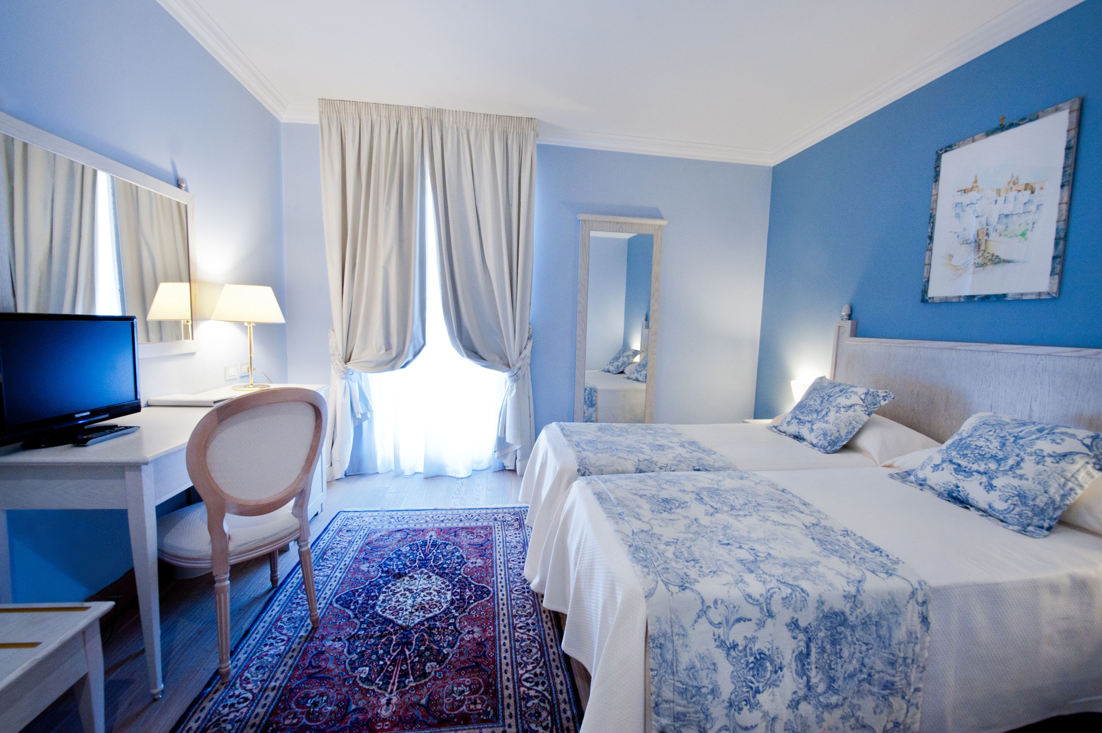 A hotel room with a bed, a desk with a television, a chair, and a large window with curtains.