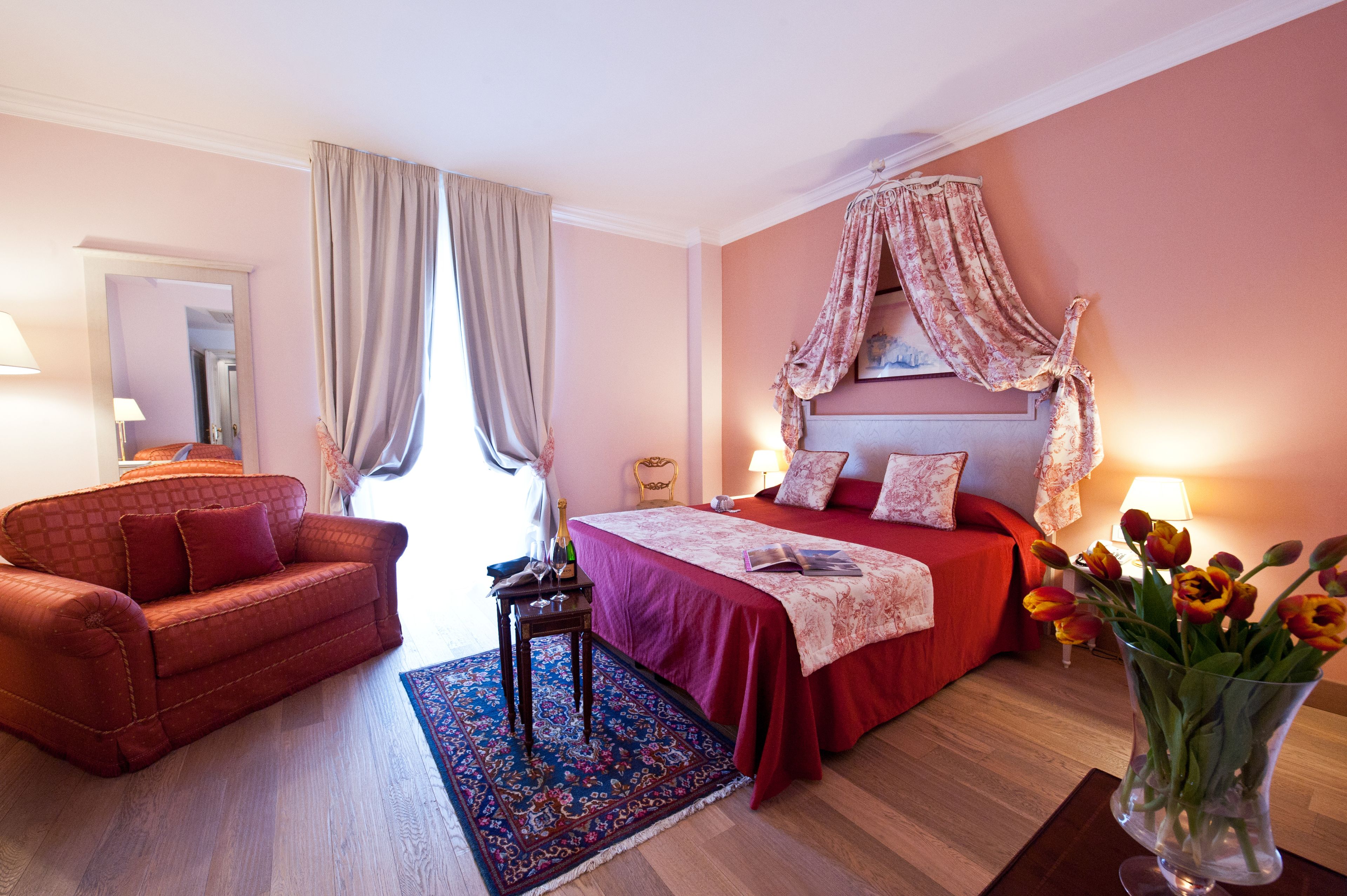Een hotelkamer met een bed, een rode bank, een klein tafeltje met een fles en glazen, een vaas met bloemen en een groot raam met gordijnen.