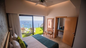 Une chambre avec un grand lit, une vue sur l’océan, un ventilateur de plafond et une porte en bois.