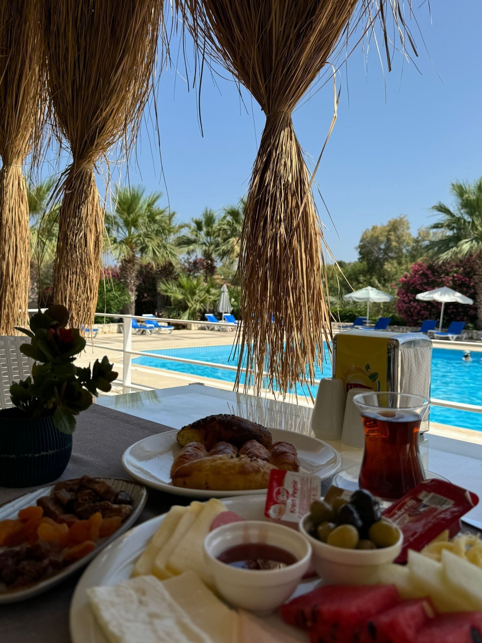 Une table dressée avec de la nourriture et des boissons, donnant sur une piscine bordée de palmiers et de chaises longues.