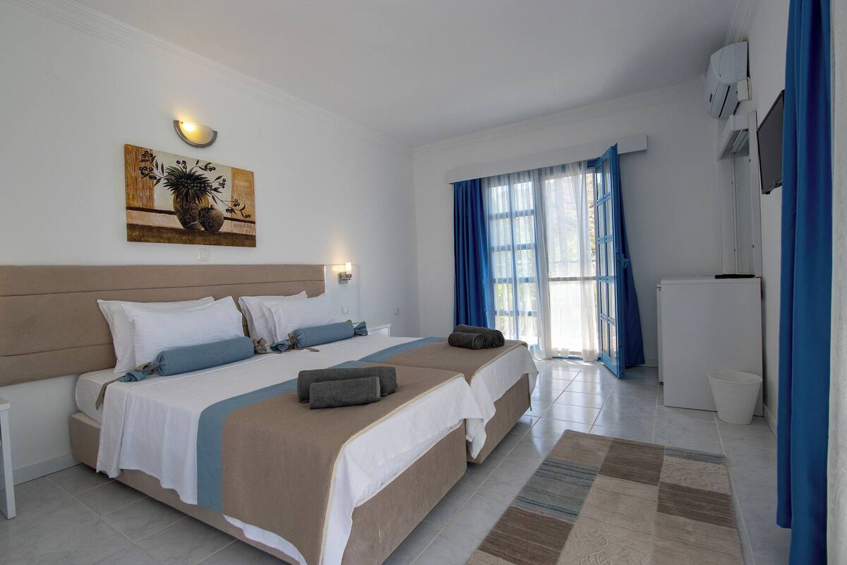 A hotel room with two beds, a large window with blue curtains, a wall-mounted air conditioner, and a small refrigerator.