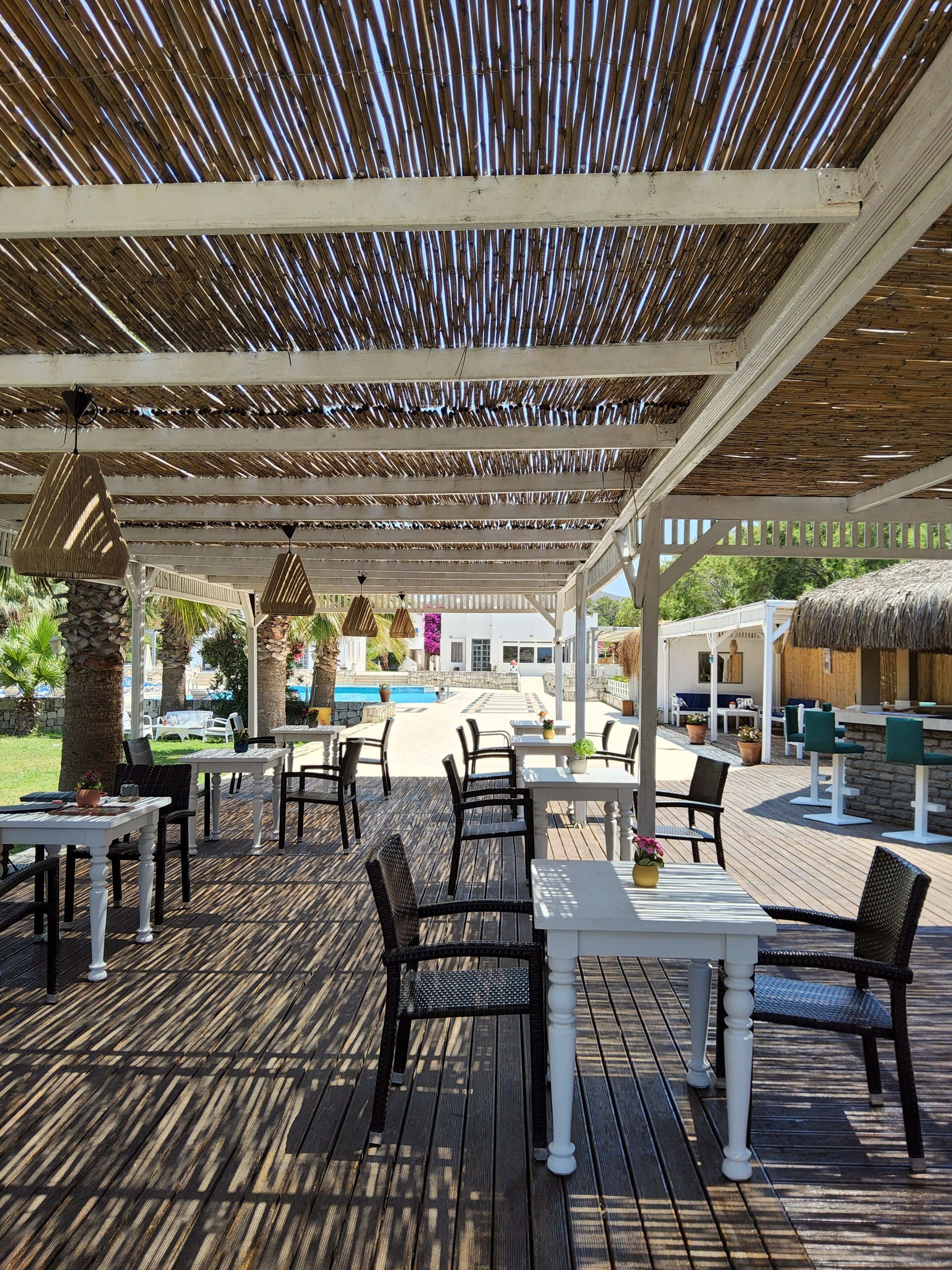 An outdoor patio with wooden tables and chairs, a pool in the background, and a thatched roof structure.
