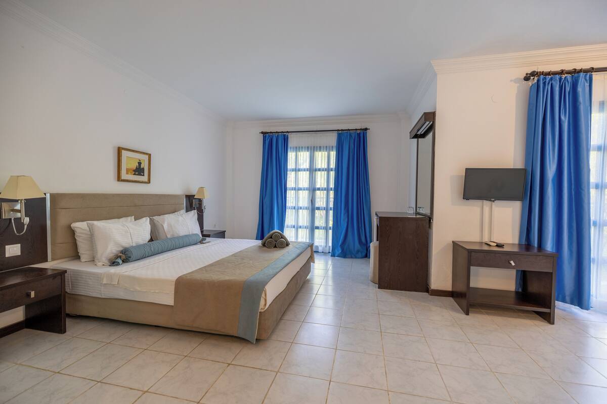 A hotel room with a bed, a bedside table with a lamp, a TV, and a desk with a chair.