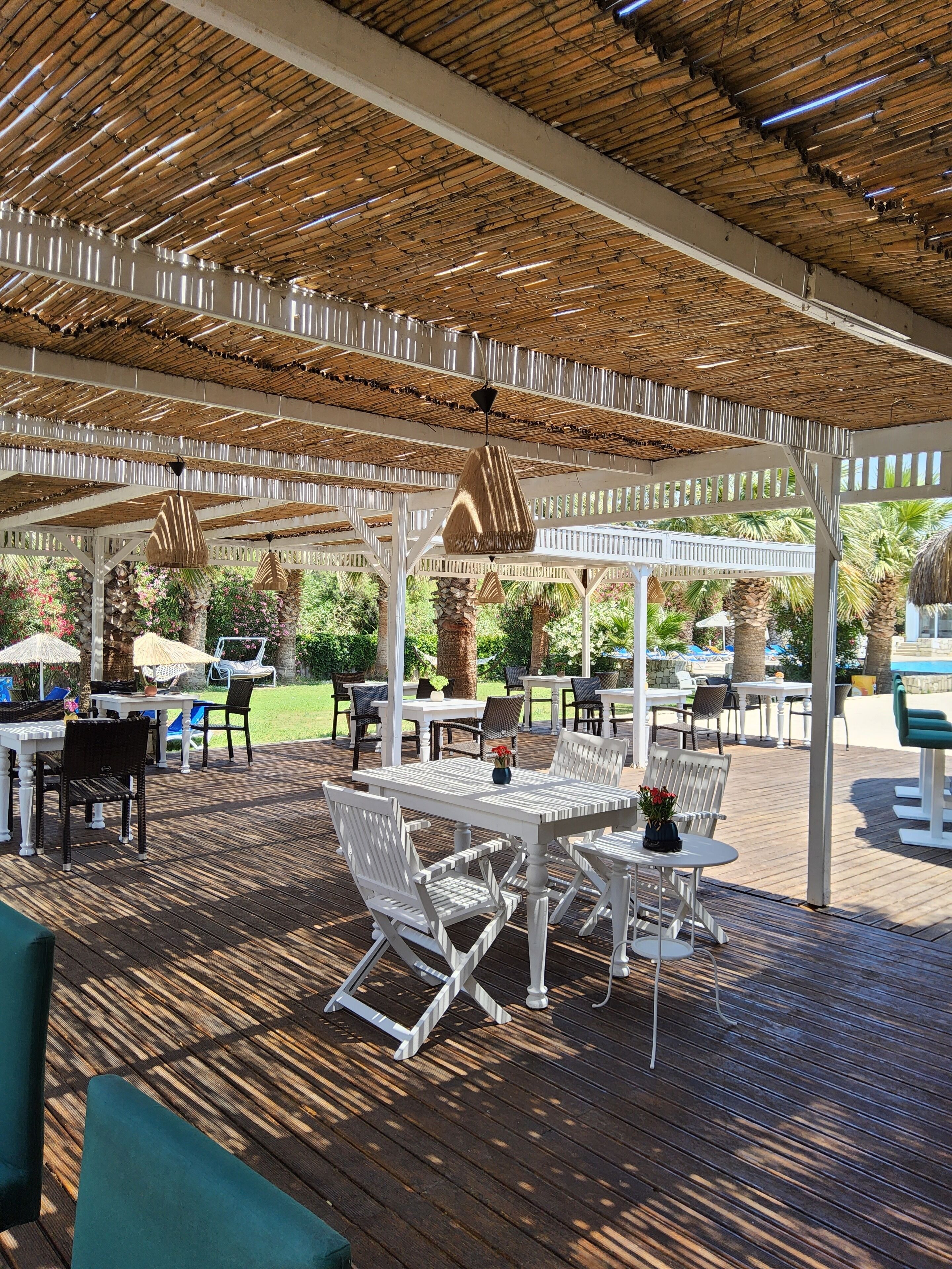 Un patio al aire libre con mesas y sillas blancas, techo de bambú y vistas a palmeras y follaje.
