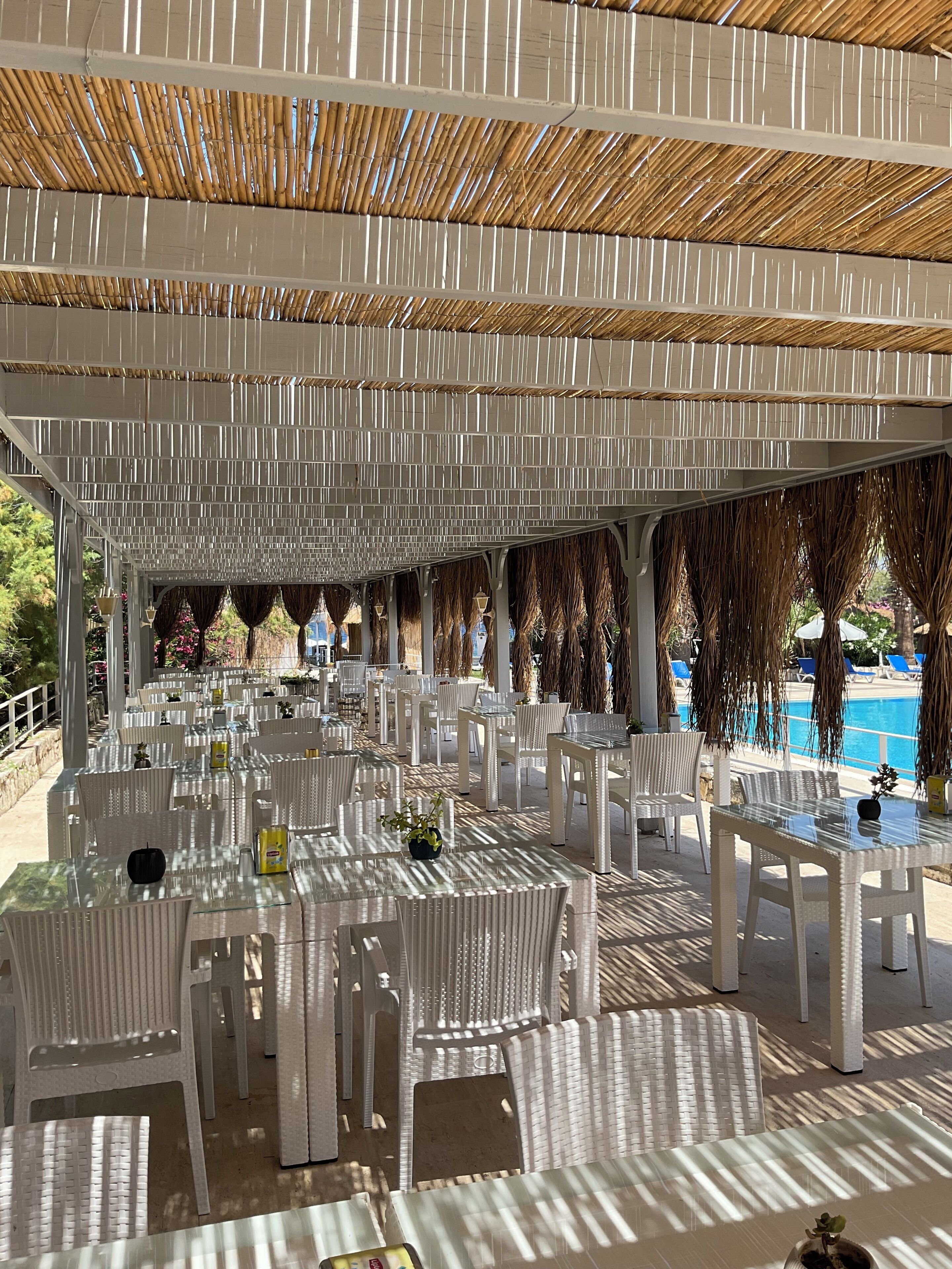 Un área de comedor al aire libre con sillas y mesas de plástico blanco, una piscina al fondo y un techo de bambú.