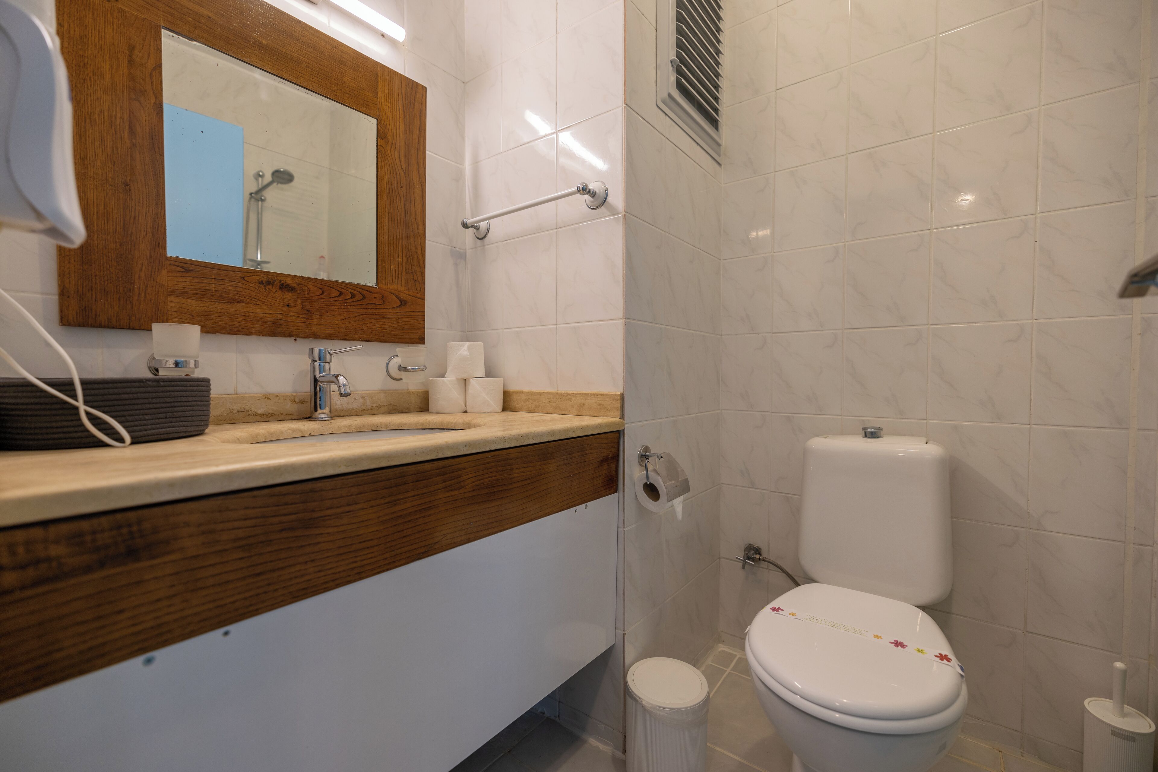 A bathroom with a wooden vanity, a mirror, a toilet, and a trash bin.