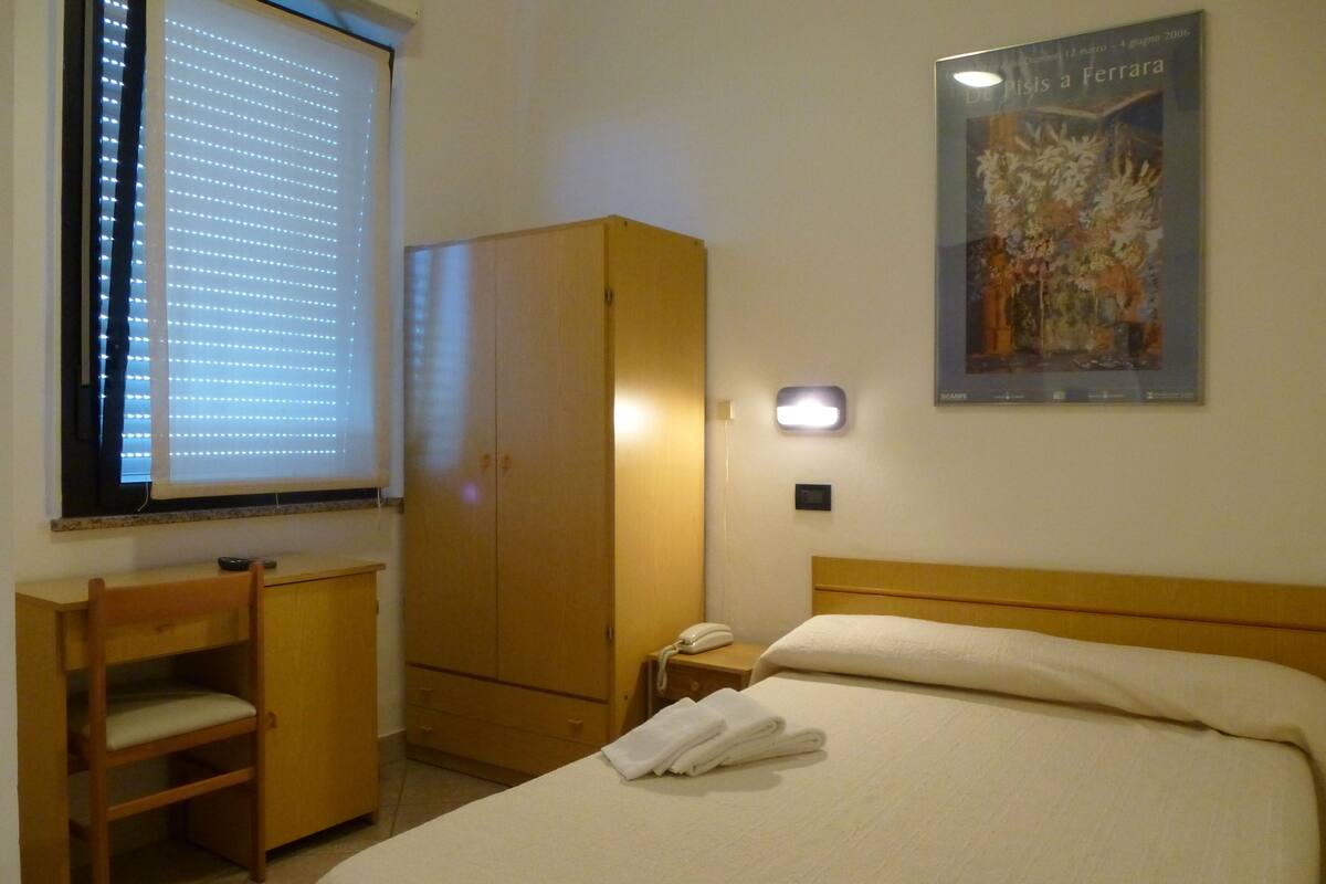 A hotel room with a bed, a wooden wardrobe, a desk with a chair, a window with blinds, and a wall-mounted light.