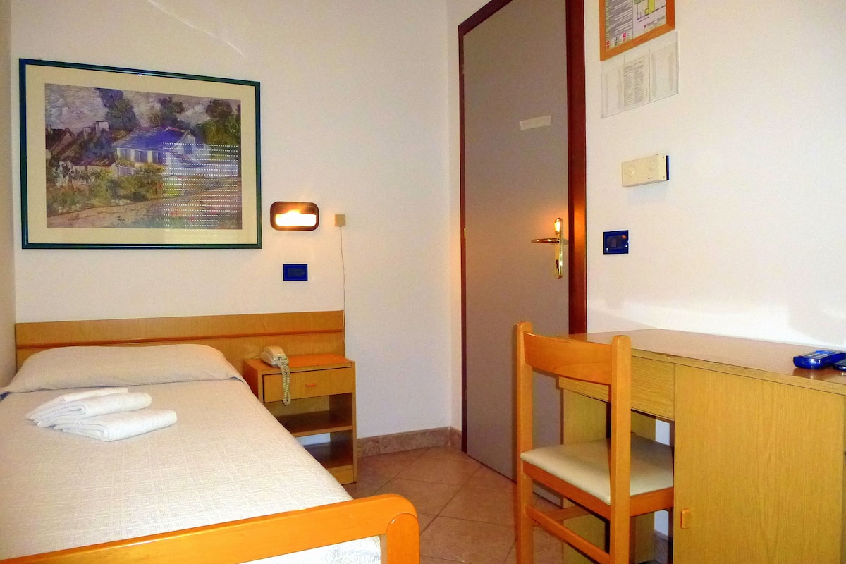 A hotel room with a bed, a desk, a chair, a bedside table, a wall-mounted telephone, and a framed picture on the wall.