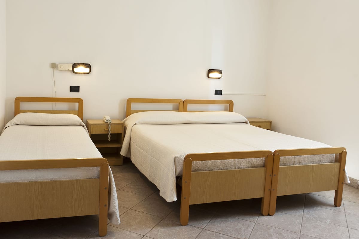 A room with two wooden beds, each with a white mattress and matching pillows. There are bedside tables with lamps and a phone on one of them. The walls are plain and white, and the floor is tiled.