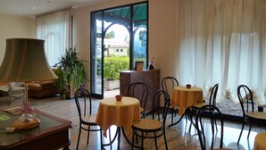 Rebedor d'hotel amb taules rodones, cadires, una làmpada i un gran finestral amb cortines.