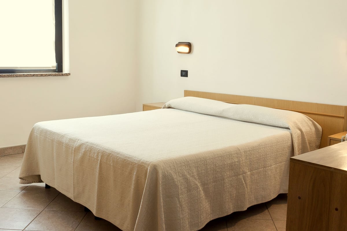 A neatly made bed with a beige bedspread in a room with a wooden headboard and a small wooden nightstand beside it.