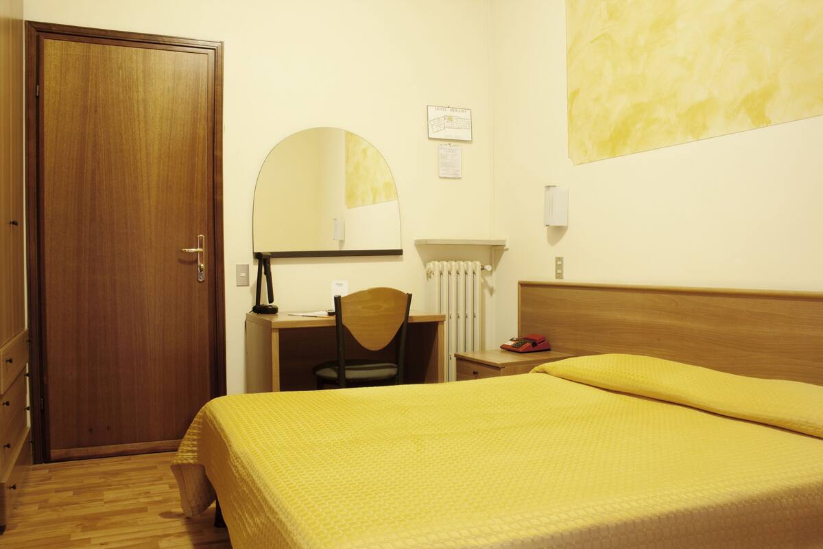 A hotel room with a wooden door, a desk with a chair, a bed with a yellow bedspread, a round mirror, and a radiator.