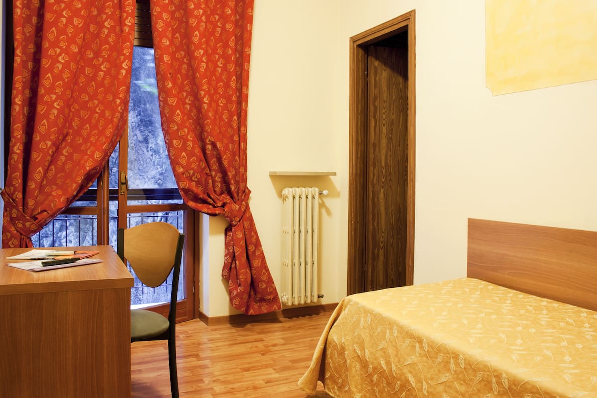 A room with a wooden desk, a chair, a bed with a patterned bedspread, a radiator, and red curtains.