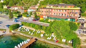 An aerial view of a coastal area with a row of colorful buildings, a dock with sun loungers, and a clear view of the water. - Hotel Merano (Brenzone sul Garda)