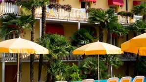 A hotel with a pool area featuring sun loungers and umbrellas, surrounded by palm trees and balconies with flower boxes. - Hotel Capri (Bardolino)