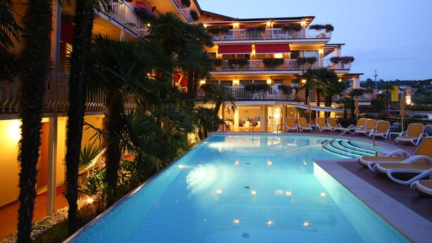 A hotel with a swimming pool, multiple balconies with plants, and outdoor seating. - Hotel Capri (Bardolino)