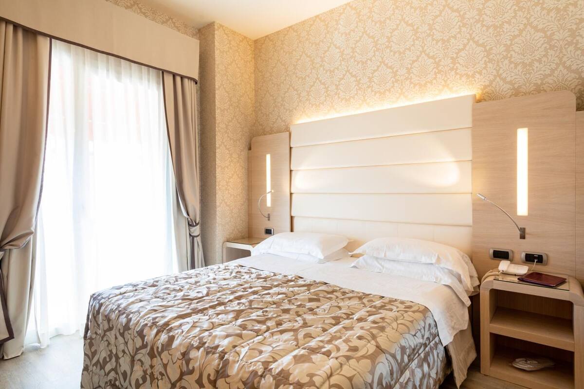 A hotel room with a large bed, a nightstand with a lamp, a bedside table with a phone, and a window with curtains.