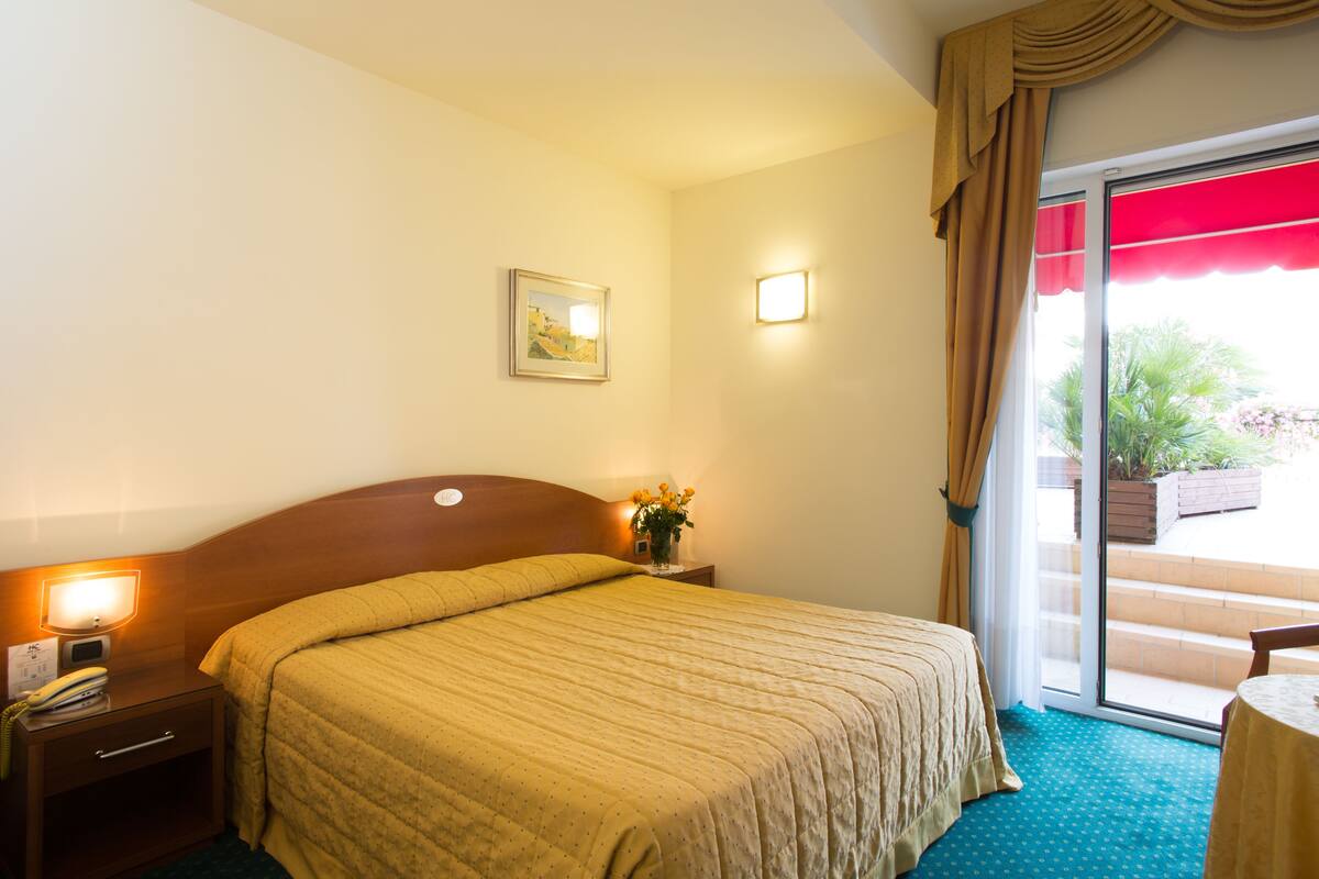 A hotel room with a bed, bedside table, lamp, and a view of the outdoors through a sliding door.