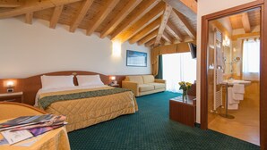 A hotel room with a wooden ceiling, a bed with a quilted bedspread, a sofa, a small table with a vase, and a bathroom visible through an open door. - Hotel Capri (Bardolino)