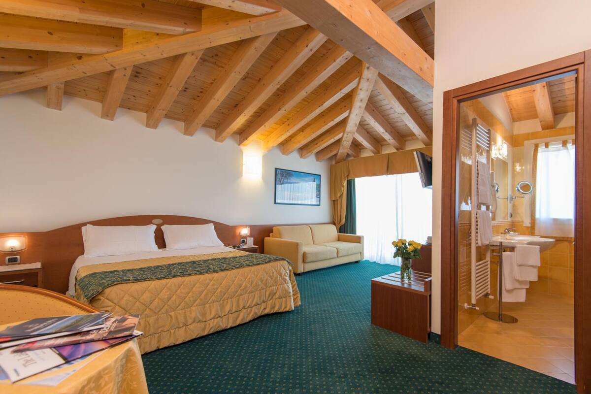 A hotel room with a wooden ceiling, a bed with a quilted bedspread, a sofa, a small table with a vase, and a bathroom visible through an open door.