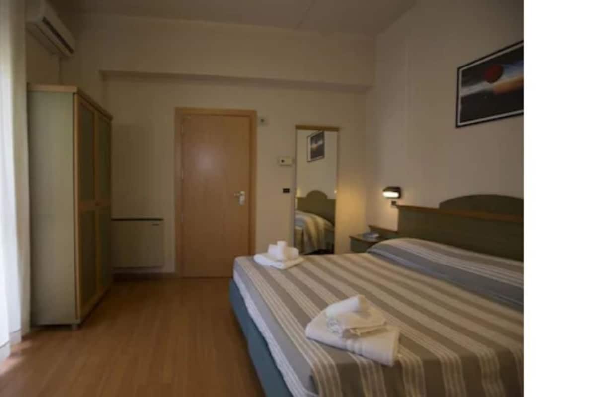 A narrow hallway leading to a bedroom with a bed, a nightstand, and a wall-mounted lamp.