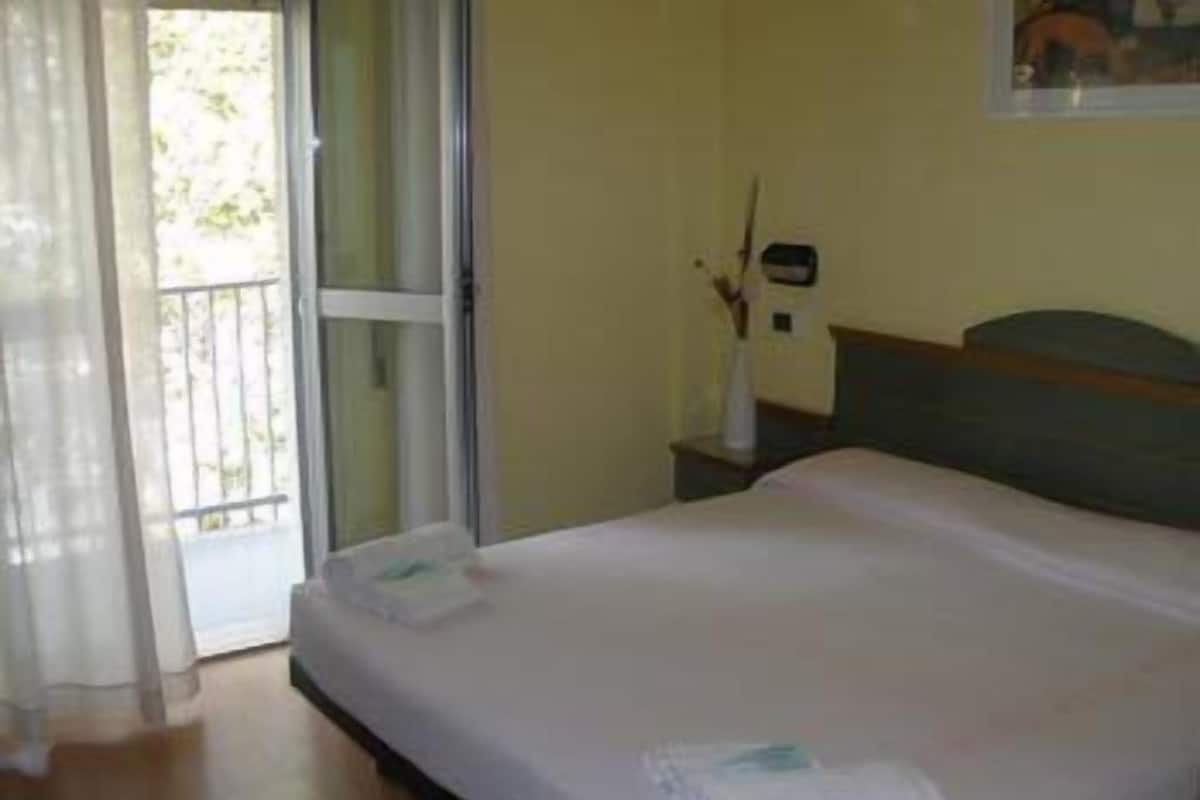 A hotel room with a bed, a nightstand, a window with curtains, and a view of greenery outside.