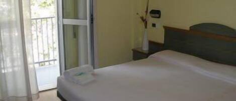 A hotel room with a bed, a nightstand, a window with curtains, and a view of greenery outside.