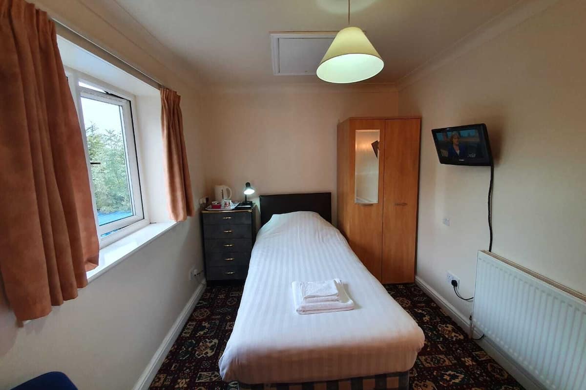Habitación de hotel con una cama individual, un armario, una mesita con lámpara, televisión y ventana con cortinas.