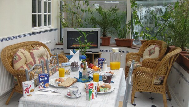 A breakfast setup with wicker chairs, a table with breakfast items, a television, and potted plants. - The Handforth Lodge (Wilmslow)