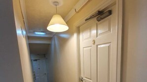 A hallway with a patterned carpet, a white door with a number plate, and a hanging light fixture. - The Handforth Lodge (Wilmslow)
