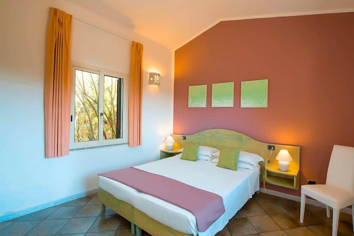 A hotel room with a bed, a chair, a window with curtains, and wall-mounted lights.