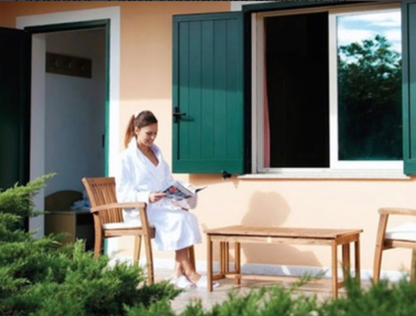 Une personne vêtue d’une robe blanche est assise à une table et à une chaise en bois, à l’extérieur, avec un bâtiment en arrière-plan.
