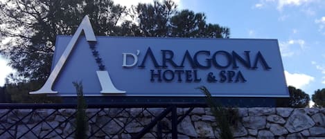 Señal del Hotel & Spa D'Aragona con un gran logotipo 'A', sobre un muro de piedra y árboles.