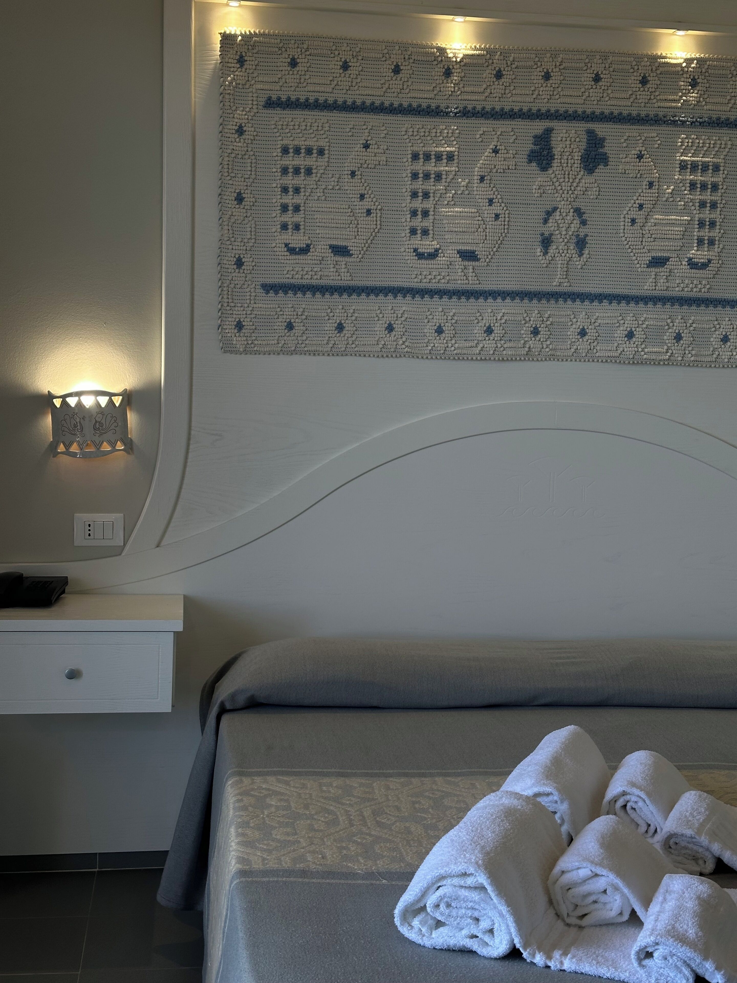 A neatly made bed with folded towels, a bedside table with a lamp, and a wall-mounted decorative piece.