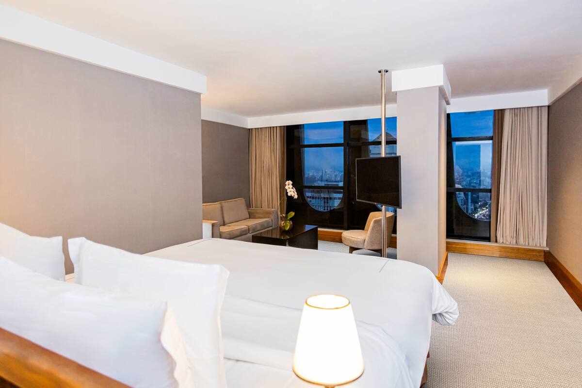 A hotel room with a large bed, a desk with a chair, a TV, and a view of the city through large windows.