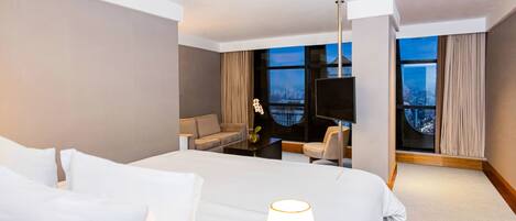 Habitación de hotel con una cama grande, un escritorio con silla, televisión y vistas a la ciudad a través de amplias ventanas.
