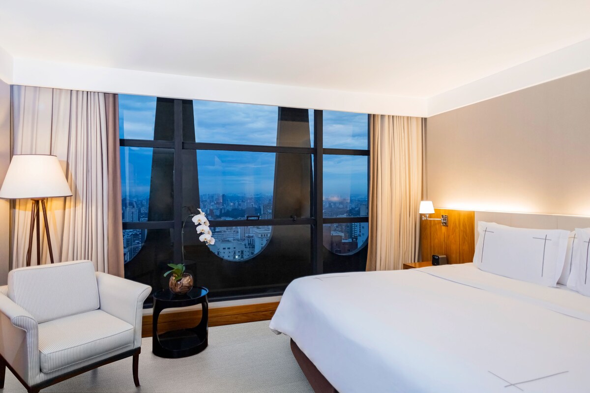 Una habitación de hotel con un ventanal grande, una cama, una silla y una pequeña mesa con una lámpara y una planta.