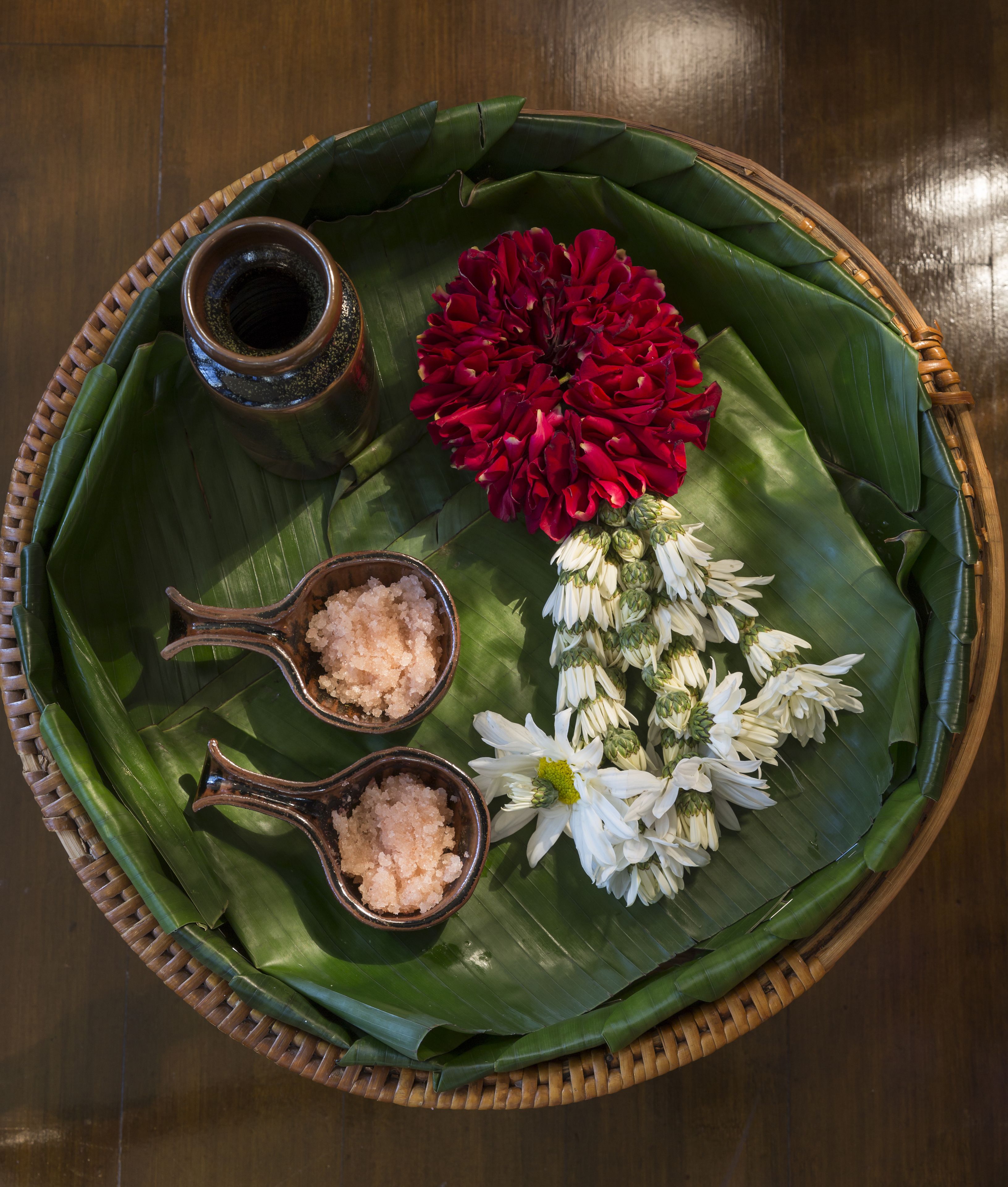 A woven basket with a green leaf base, containing two small bowls of pink salt, a red flower, and a bunch of white flowers.
