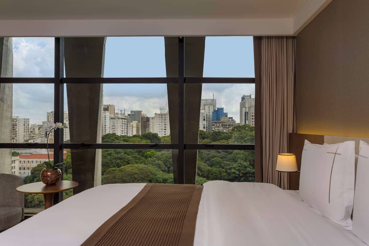 A hotel room with a large window, a bed, a bedside table with a vase, and a lamp.