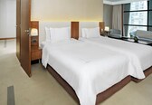 A hotel room with two beds, a nightstand with a lamp, a chair, and a view of the city through large windows.